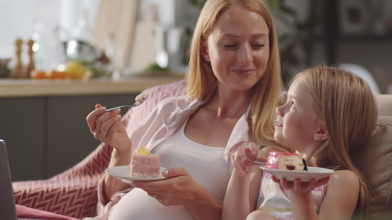 chica feliz comiendo postre y viendo una película con su madre embarazada