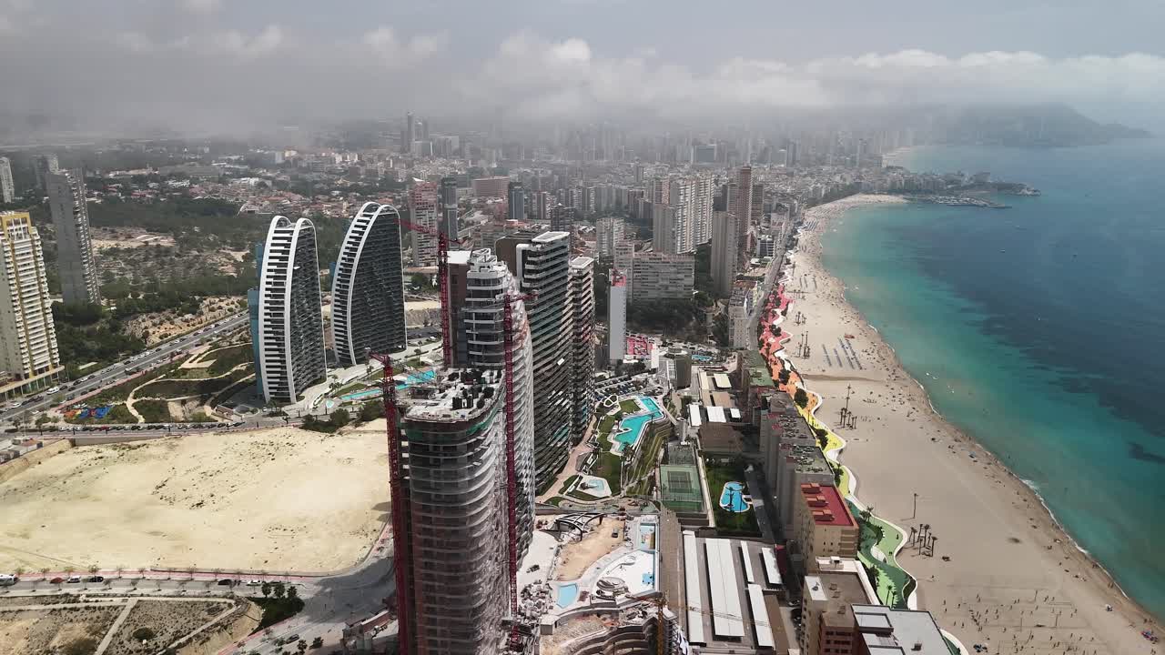 Aerial pass over Benidorm, Spain: Drone glides, then camera tilts to zenithal view above this famed resort's skyscrapers, beach, new builds, and iconic skyline. Alicante.
