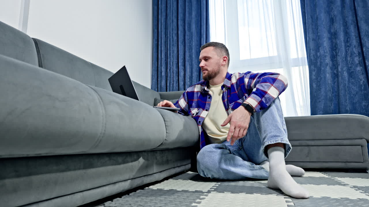 Hombre sentado en el suelo y trabajando en un portátil en la sala de estar de día en un apartamento moderno con ventanas