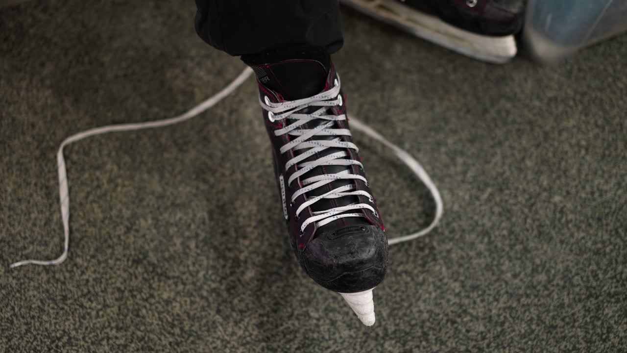 Close-up view of a black ice skate with red accents being tied around the leg, the person is wearing black trousers and a brown sleeve top