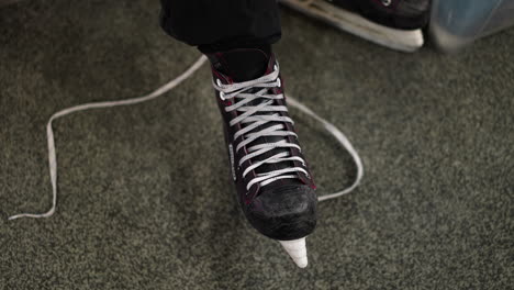 close-up view of a black ice skate with red accents being tied around the leg, the person is wearing black trousers and a brown sleeve top