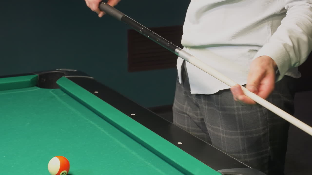 Person in white shirt and grey trousers, face not visible, picking up cue stick beside green pool table. Focus on hands, billiard balls, and equipment. Professional game setting