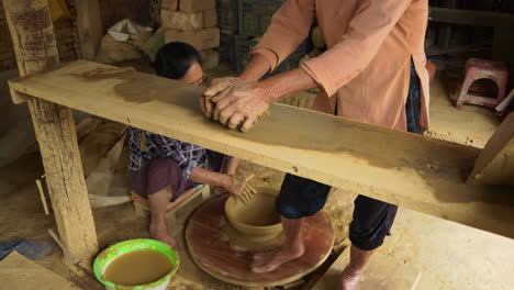 two asian women make traditional pottery: one kneads new clay, and the other finishes a big bowl at a turning table powered by foot