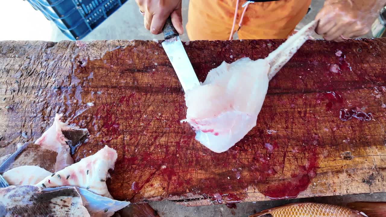 Närbild på en fiskhandlare som filéar färsk fisk på en rustik träskärbräda, vilket visar beredning av råa skaldjur i en utomhusmarknadsmiljö. Händer använder kniv för att skära vitt fiskkött