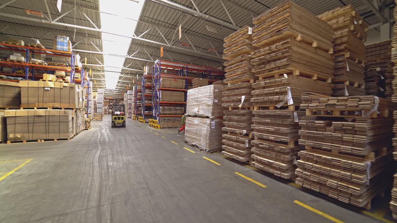 Operator in a helmet drives a yellow electric forklift through the warehouse in search of the right product on the background of huge shelves.