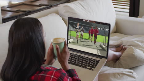 compuesto de una mujer sentada en casa sosteniendo un café viendo un partido de rugby en una computadora portátil