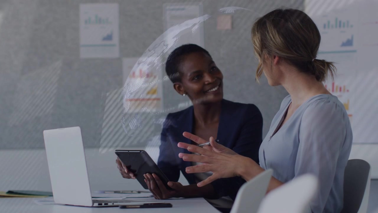 Two women at meeting table, tapping tablet and initiating faint globe overlay, discussing business