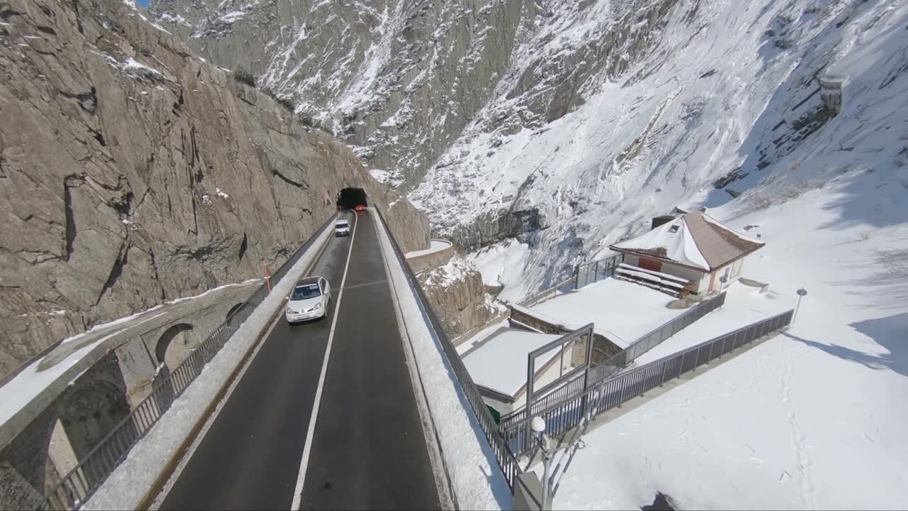 Cars travelling over a mountain pass with snow, rocks, cliffs and valleys and the roof of some buildings