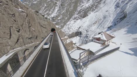 cars travelling over a mountain pass with snow, rocks, cliffs and valleys and the roof of some buildings