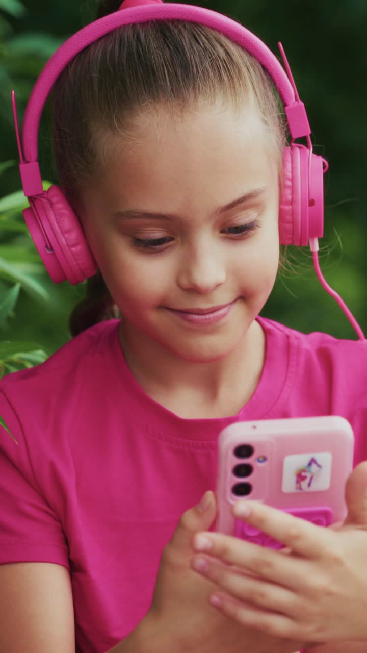 A Joyful Moment: A Young Girl Enjoying Music with Pink Headphones and a Smartphone in a Lush Green Outdoor Setting