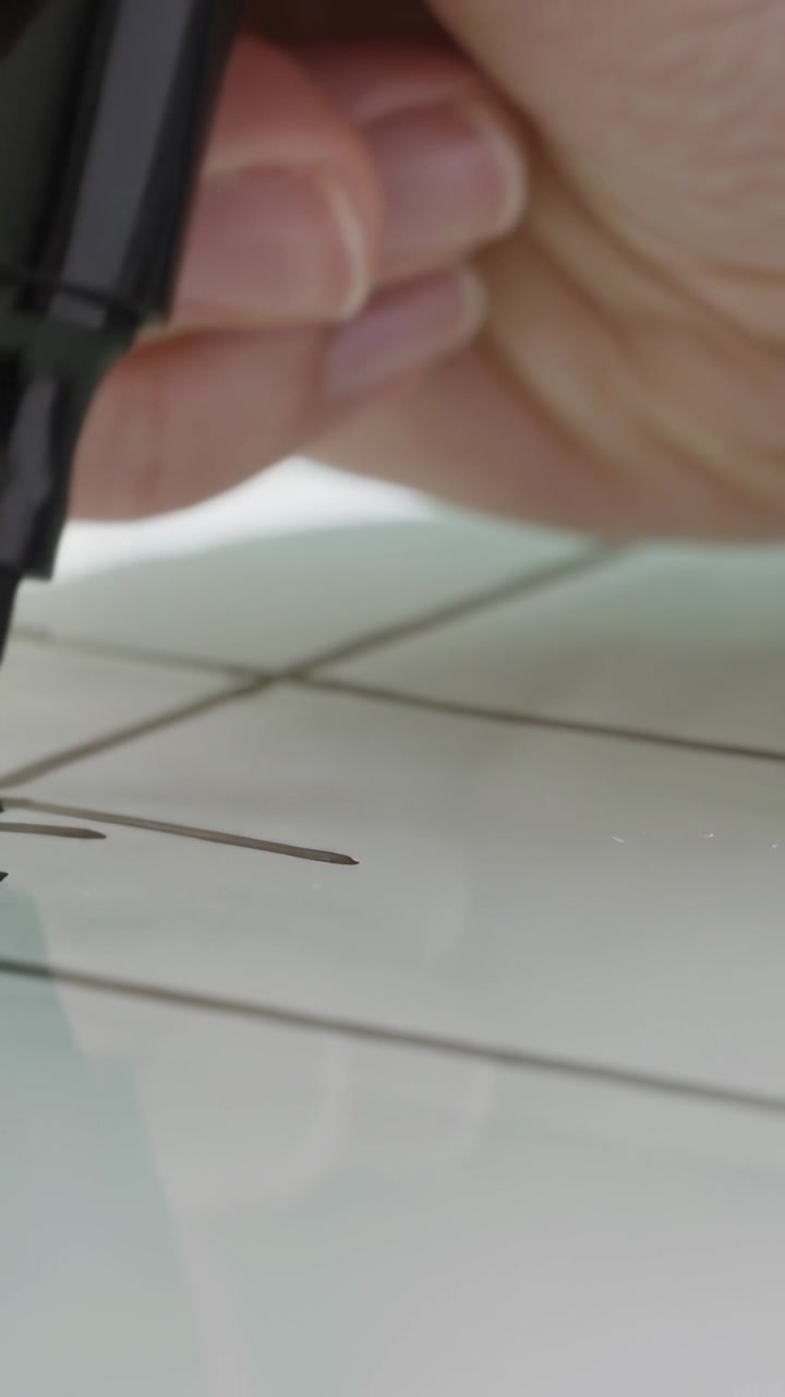 Vertical video: Lowering adult hand drawing crossing lines on glossy grid panel, with black marker