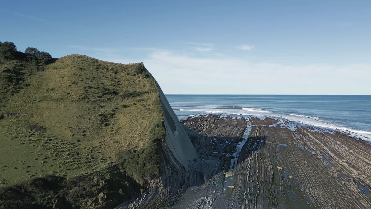 Fremoverbevegende luftfoto som viser de spektakulære flysch-fjellformasjonene på den baskiske kysten ved Sakoneta-stranden, nær Zumaia, og avslører kystlinjens robuste skjønnhet under en klar blå himmel.