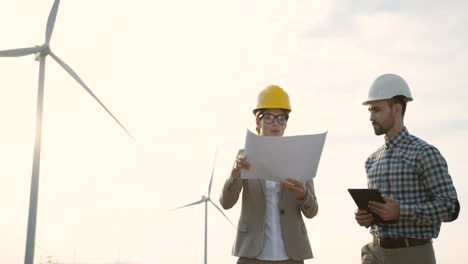 Caucasian-man-and-woman-engineers-wearing-a-helmet-watching-some-blueprints-and-using-tablet-while-talking-at-wind-station-of-renewable-energy