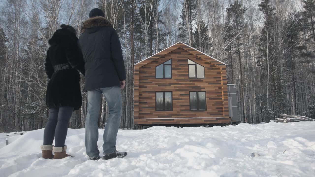 una pareja mirando una nueva cabaña de madera en un bosque nevado