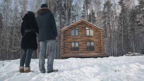 una pareja mirando una nueva cabaña de madera en un bosque nevado