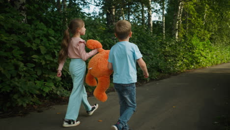 rear view of two siblings walking side by side on quiet paved path surrounded by dense greenery, with girl gently lifting large orange teddy bear off ground while walking beside younger brother