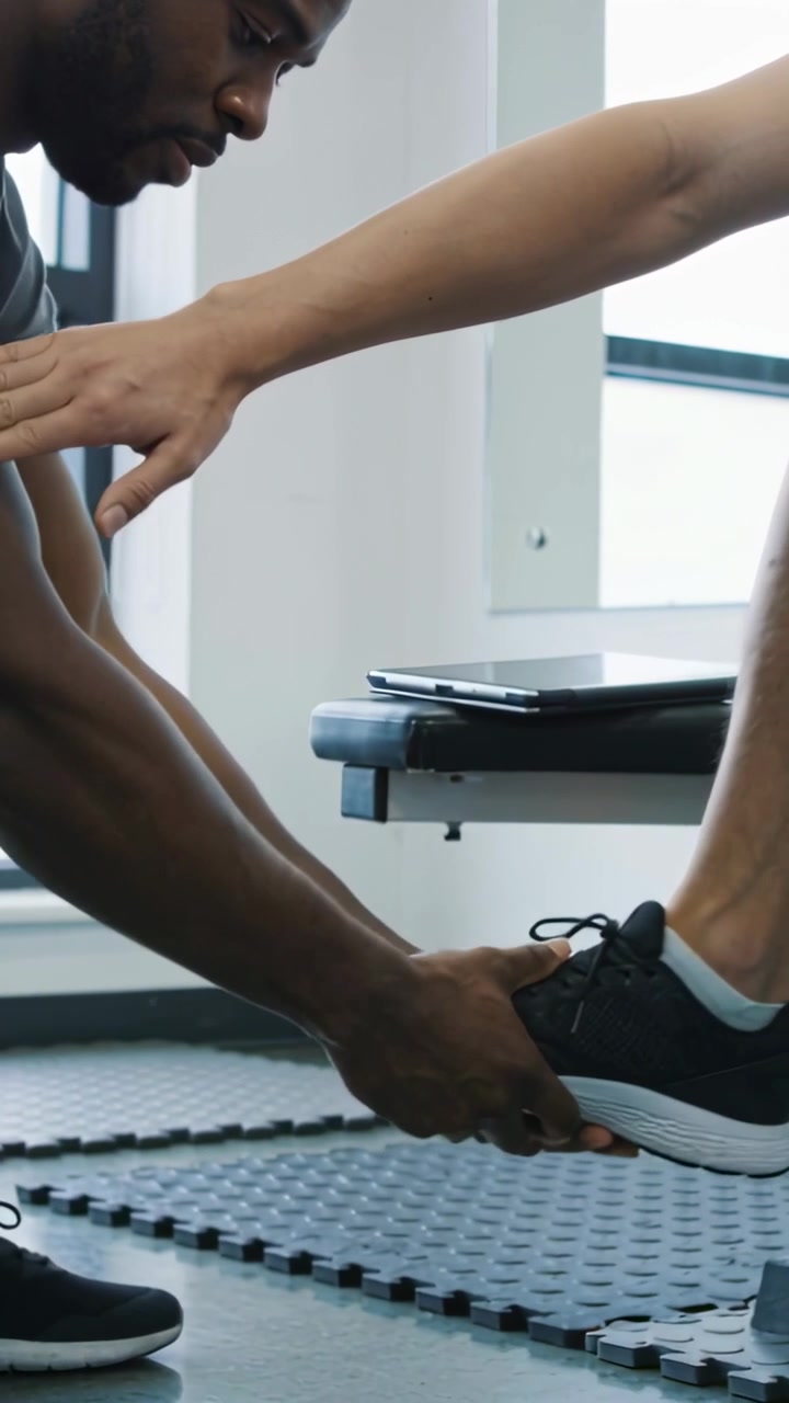 Vertical video: Kneeling trainer fixing sneaker at bench with client holding arm out, copy space