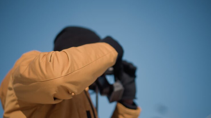 back view of operator in yellow jacket and knit beanie peering through vintage camera viewfinder gloved hands adjusting focus dial under clear urban winter sky bright sunshine over snowy street