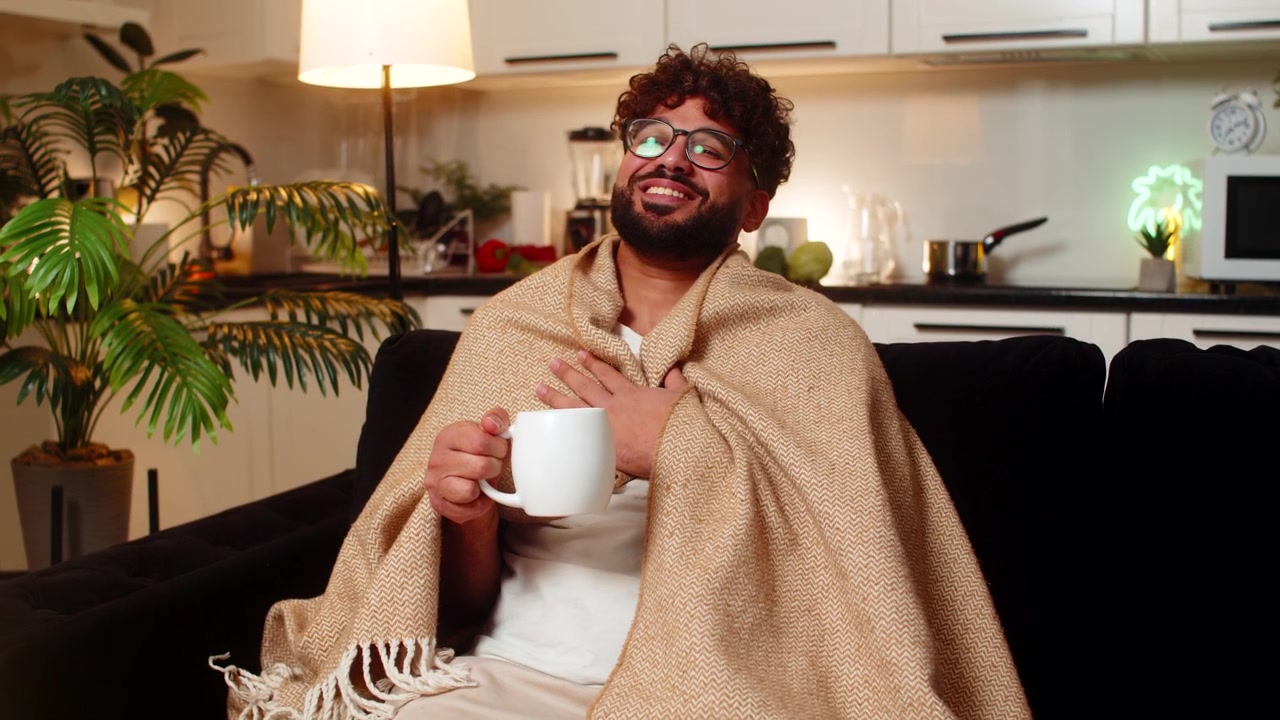 Uomo arabo avvolto in una coperta che beve tè caldo e si scalda le mani con la tazza, in una pausa lenta in una serata calma e accogliente.