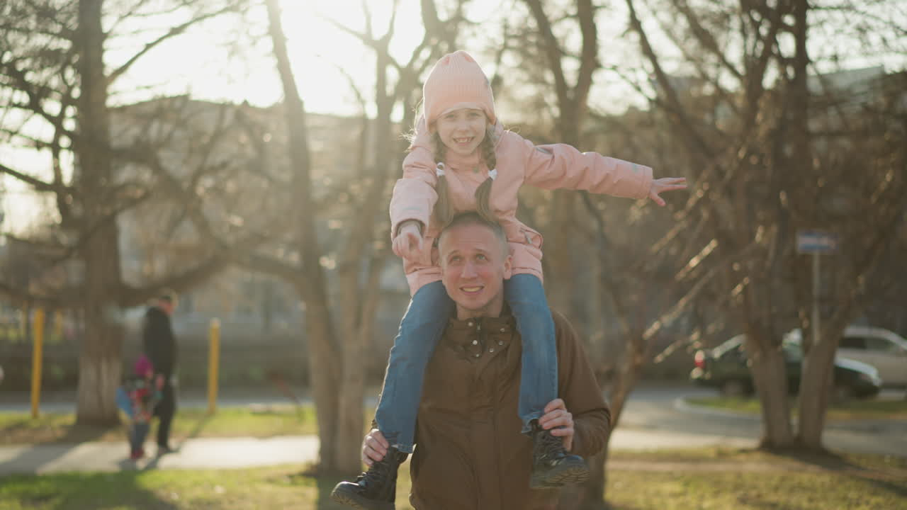 A joyful little girl in a pink cap and jacket spreads her arms wide while sitting on her father's neck as they walk through a sunlit park. Her father, wearing a brown jacket
