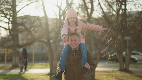 a joyful little girl in a pink cap and jacket spreads her arms wide while sitting on her father's neck as they walk through a sunlit park. her father, wearing a brown jacket