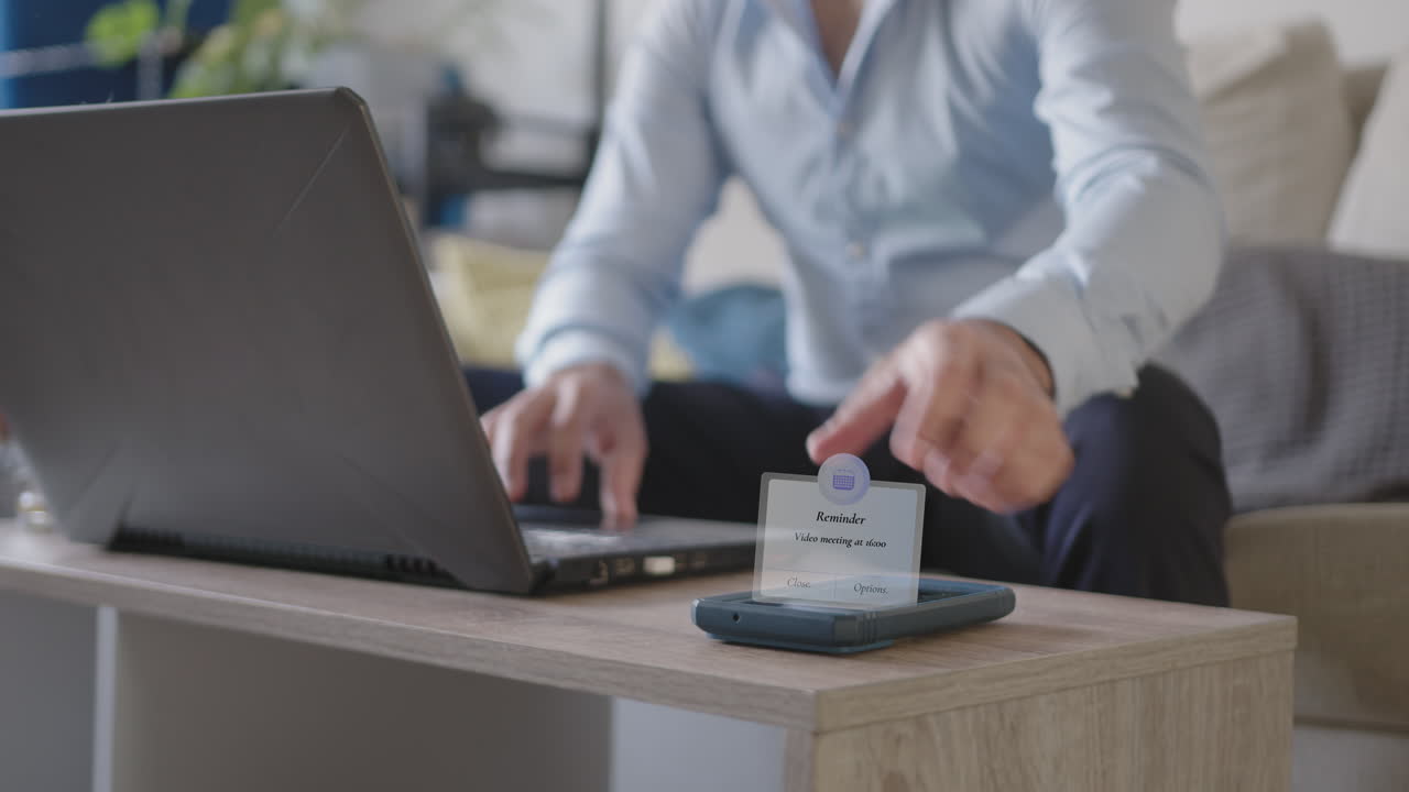 teléfono móvil video reunión recordatorio popup distracción joven adulto freelance hombre de negocios trabajando desde casa en su sala de estar usando portátil, con una camisa de botones azul claro