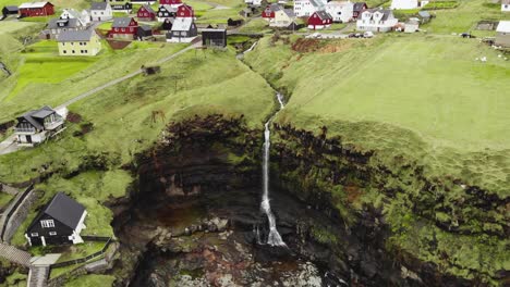 Antenne-Eines-Kleinen-Dorfes-Auf-Der-Klippe-Mit-üppigem-Gras-Und-Kleinen-Flüssen-Und-Wasserfällen,-Die-In-Den-Ruhigen-Nordatlantik-Im-Fjord-Auf-Den-Färöer-Inseln-Fallen