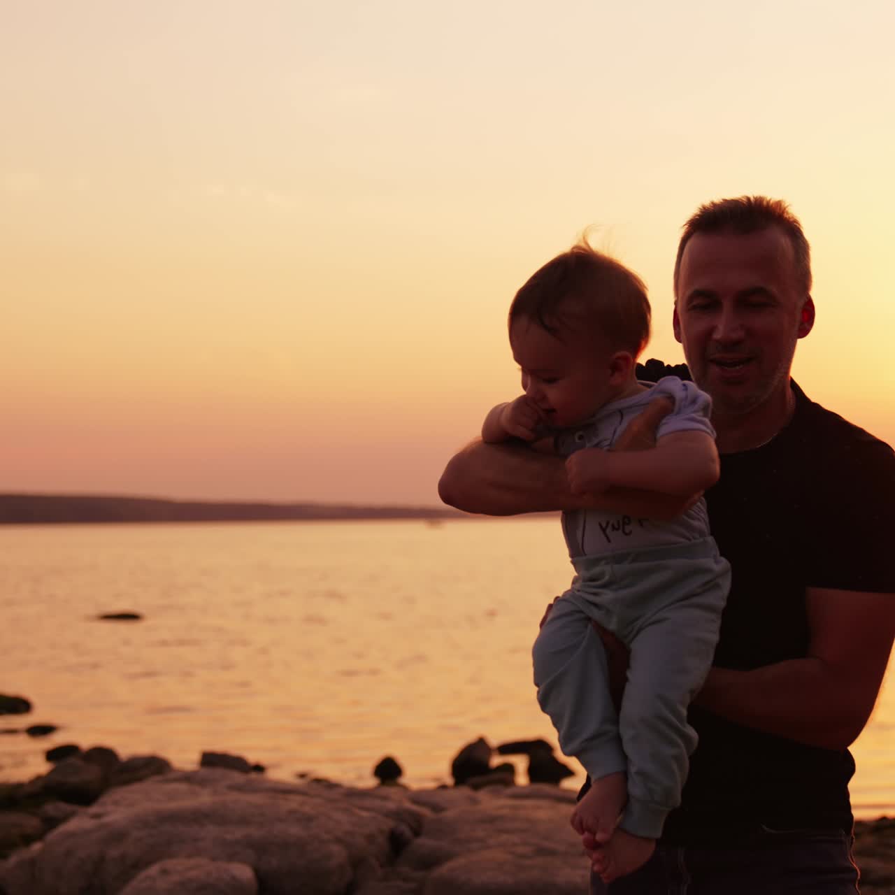 Padre affettuoso che trascorre del tempo con il suo bambino. Il padre tiene in braccio suo figlio, lo fa girare e lo lancia in aria. Tramonto sul fiume sullo sfondo