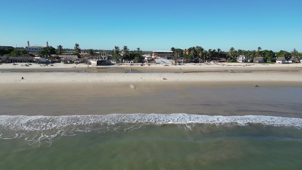 Toma aérea baja con dron a lo largo de la costa de la playa de Morondava, Madagascar. El video muestra olas golpeando suavemente el banco de arena cerca de estructuras frente al mar y casas.