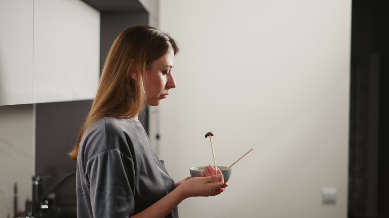 Side view of lady in casual gray shirt standing in modern kitchen using single chopstick to pick up piece of meat from bowl, enjoying quiet meal, bright white cabinet background