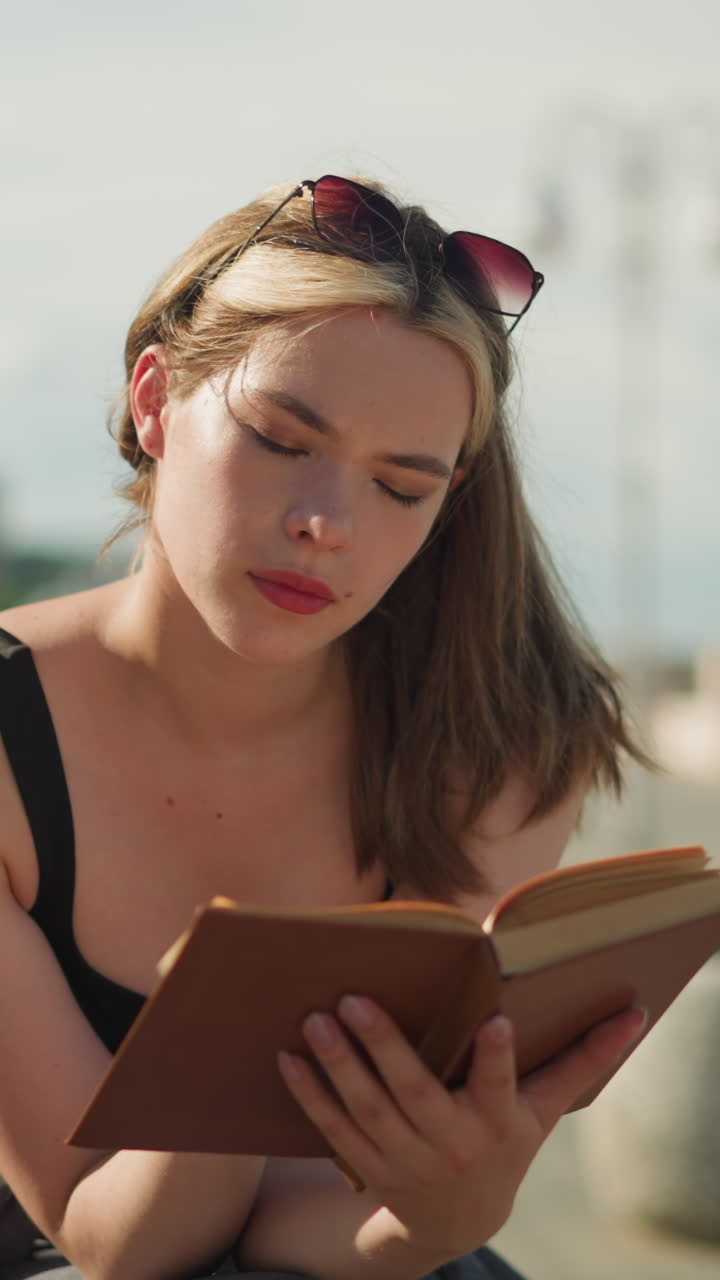 dama de soltera negra se sienta al aire libre, leyendo un libro cubierto de marrón mientras el viento voltea las páginas ligeramente, expresión pensativa en su rostro con un fondo borroso de personas que pasan