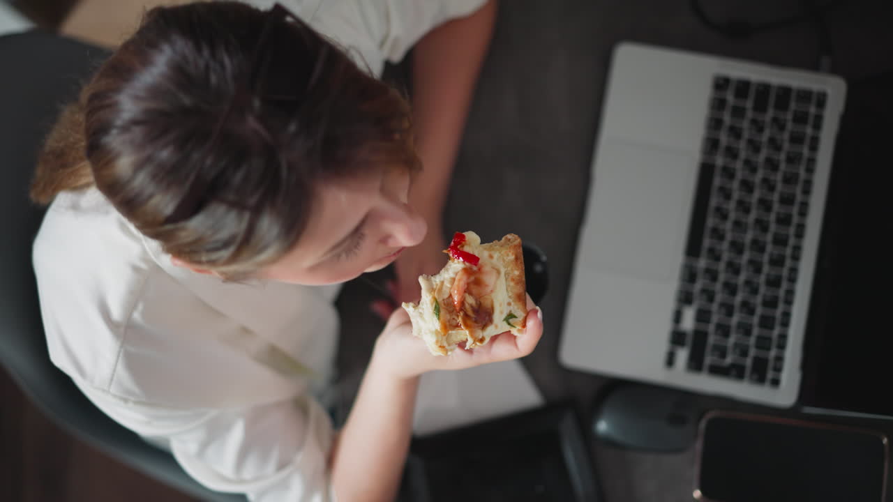 Overhead view of woman wearing white shirt sitting at desk eating delicious pizza slice with melted cheese and toppings during work break, laptop, phone, mouse, and papers scattered on dark worktable