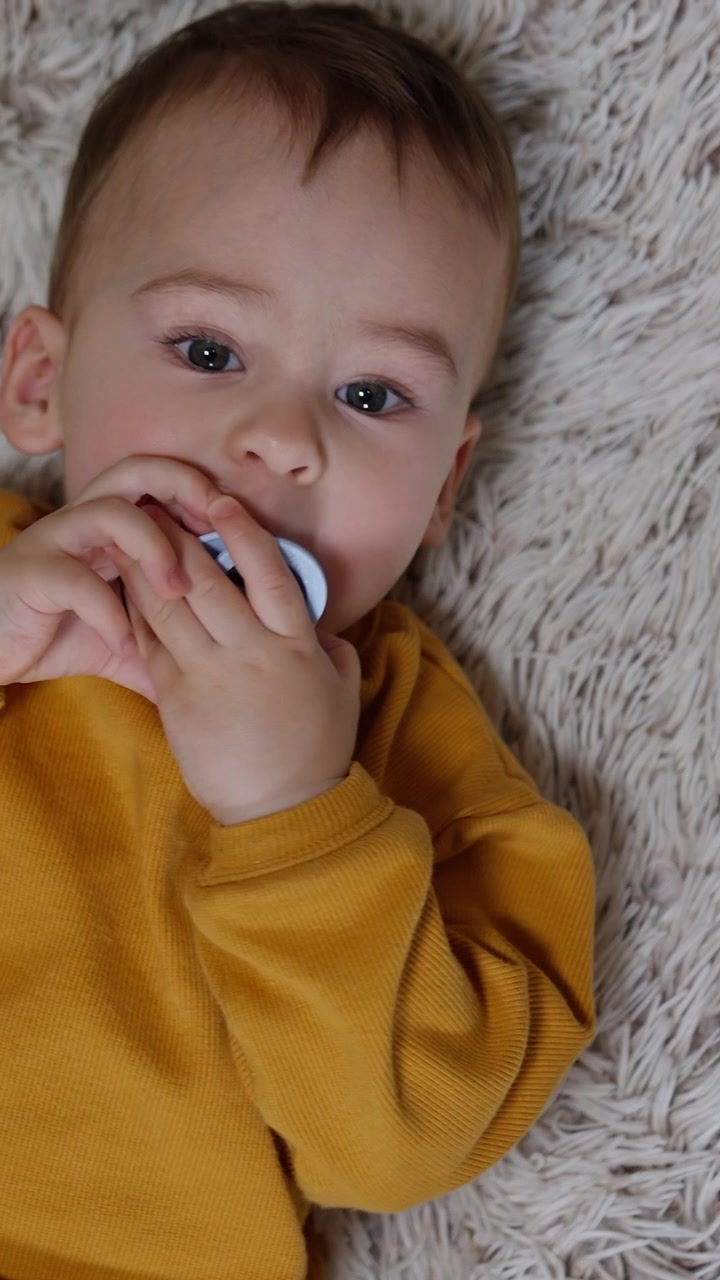 Sweet kid rolls by the soft fluffy carpet. Little boy in orange sport suit focused on the pacifier. View from top. Vertical video