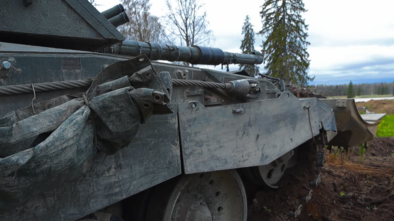 British military Challenger 2 4034 bulldozer tank with a plough at the front aiming its large cannon weapon at an open field during a cloudy day from a forest. Tank is looking for enemies in distance.