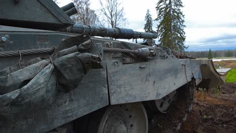 british military challenger 2 4034 bulldozer tank with a plough at the front aiming its large cannon weapon at an open field during a cloudy day from a forest. tank is looking for enemies in distance.