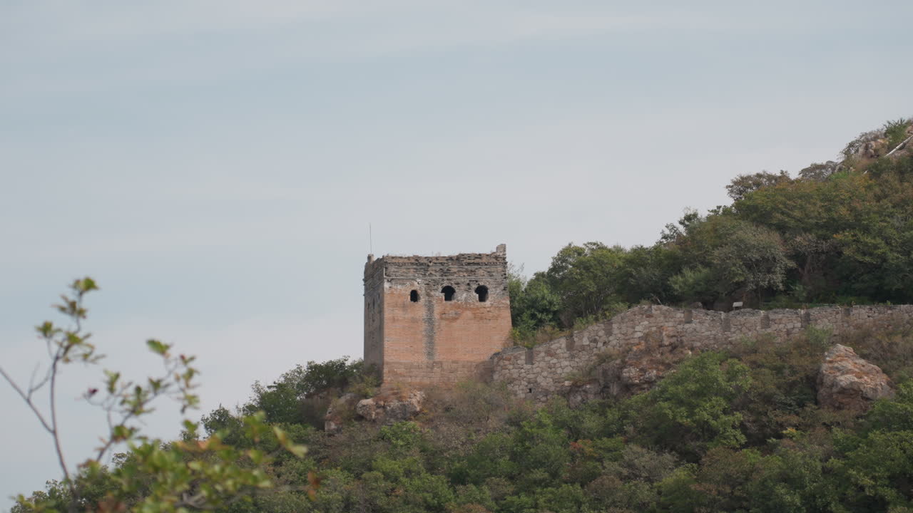 Torre de piedra solitaria en la cima de una colina, perspectiva aérea cinematográfica sobre un muro de piedra erosionado y vegetación densa, cielo tranquilo y horizonte panorámico, ambiente rústico e histórico ideal para viajes, patrimonio, documentales.