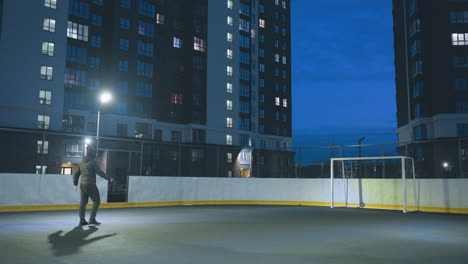 athlete kicks soccer ball toward goal during intense night practice on urban sport field surrounded by towering residential buildings and illuminated by vibrant blue evening sky and streetlights