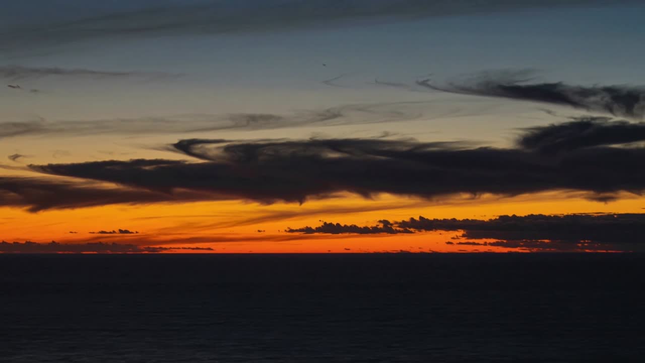 Sich verlagerndes Wolkenband bewegt sich über einen farbenfrohen Himmel über offenem Meereshorizont bei Sonnenuntergang, mit orangefarbenem Glühen.