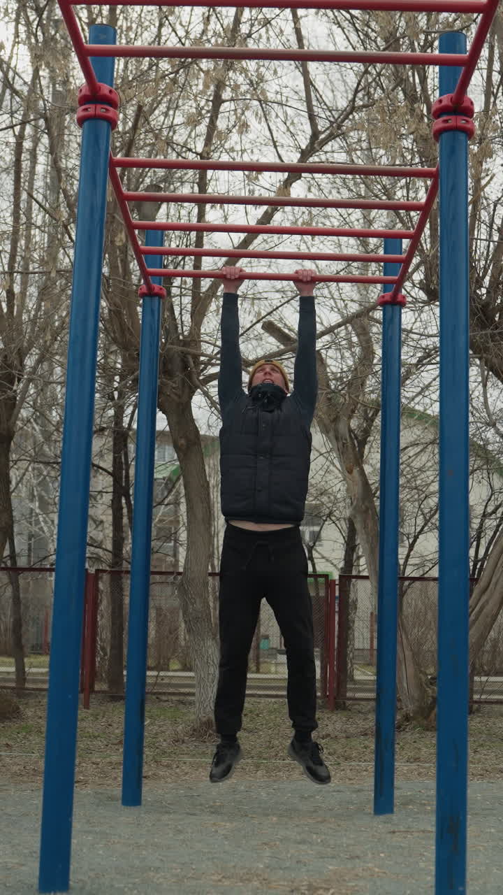 A coach jumps up and grabs a red iron bar, slowly moving across it with controlled strength, in the background, a boy plays football, and a residential building is visible across the road