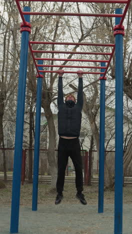 a coach jumps up and grabs a red iron bar, slowly moving across it with controlled strength, in the background, a boy plays football, and a residential building is visible across the road