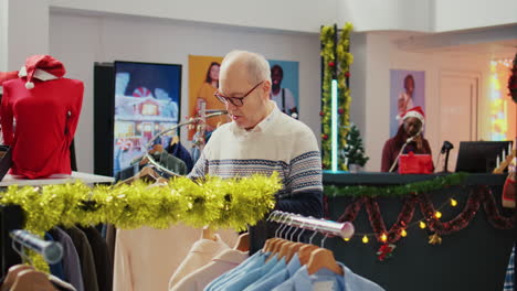 Dolly-in-shot-of-senior-customer-in-festive-ornate-shopping-mall-store-trying-to-determine-if-stylish-blazer-is-fitting-him.-Aged-client-in-fashion-shop-during-Christmas-holiday-season