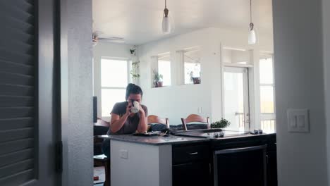 wide shot of a pretty millennial caucasian woman slowly slips a morning cup of coffee while sitting in her kitchen and dining room
