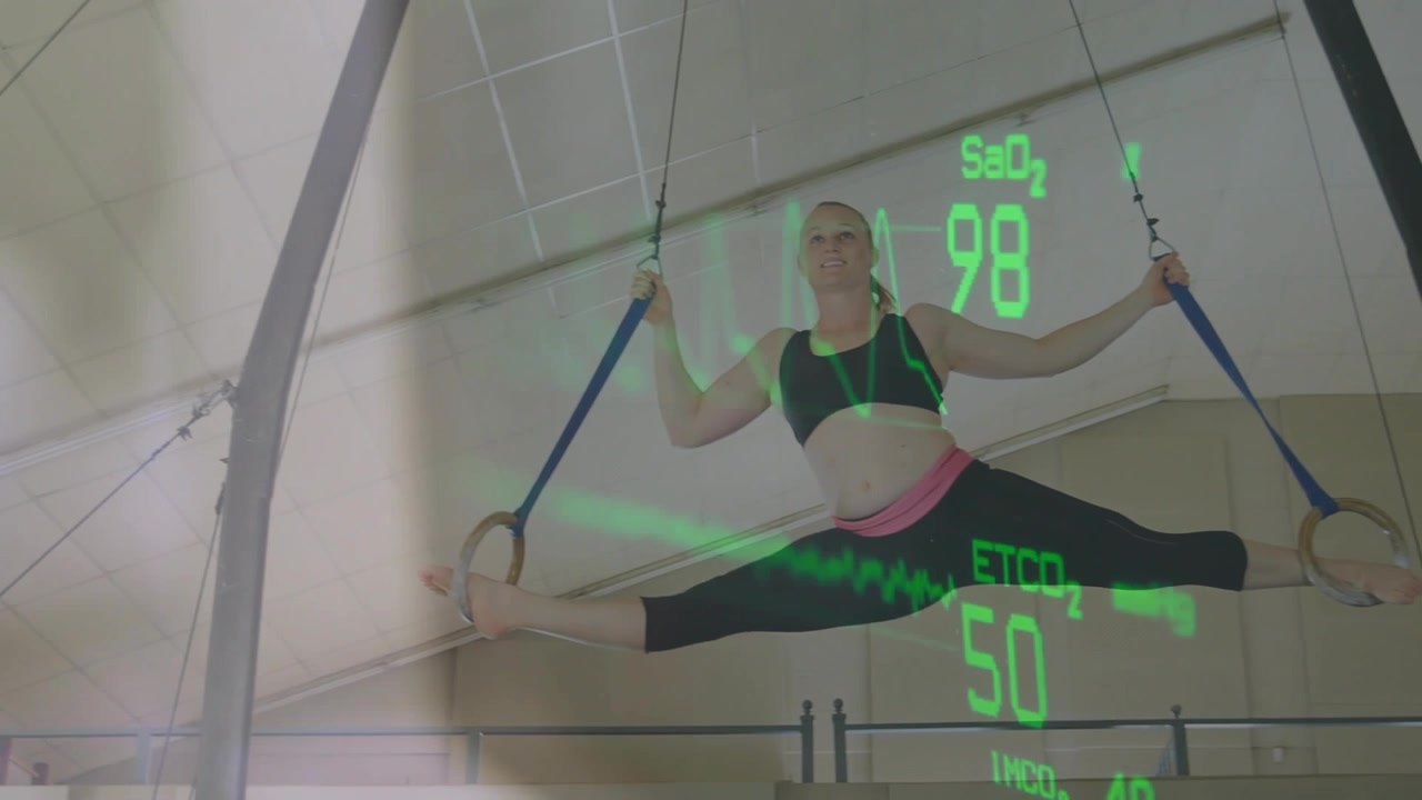 Female gymnast gripping straps, pulling into rings in gym while fitness overlay tracking SaO2 or ETC