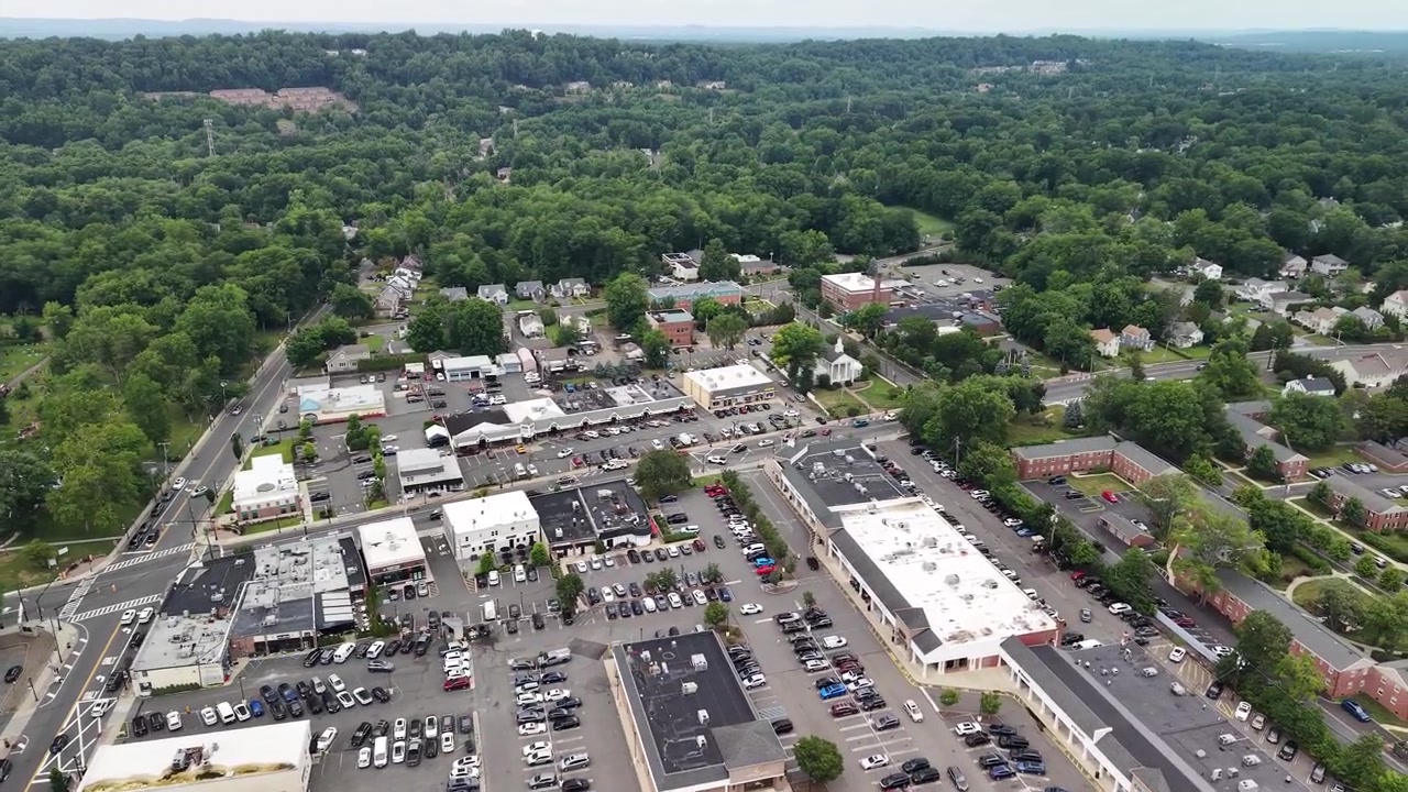 Luftperspektiv der viser en charmerende lille amerikansk by med travle gader, parkeringspladser ved indkøbscentre og kommercielle bygninger, omgivet af et stort område med frodig grøn skov