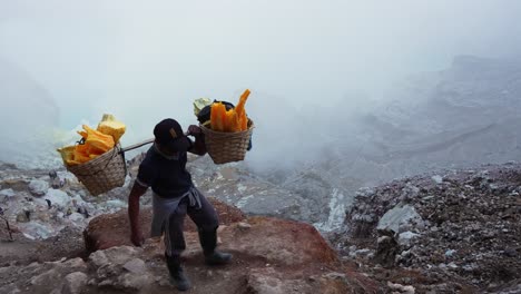 miner carrying extremely heavy sulfur load out of kawah ijen volcano crater during the day in east java, indonesia, wide shot