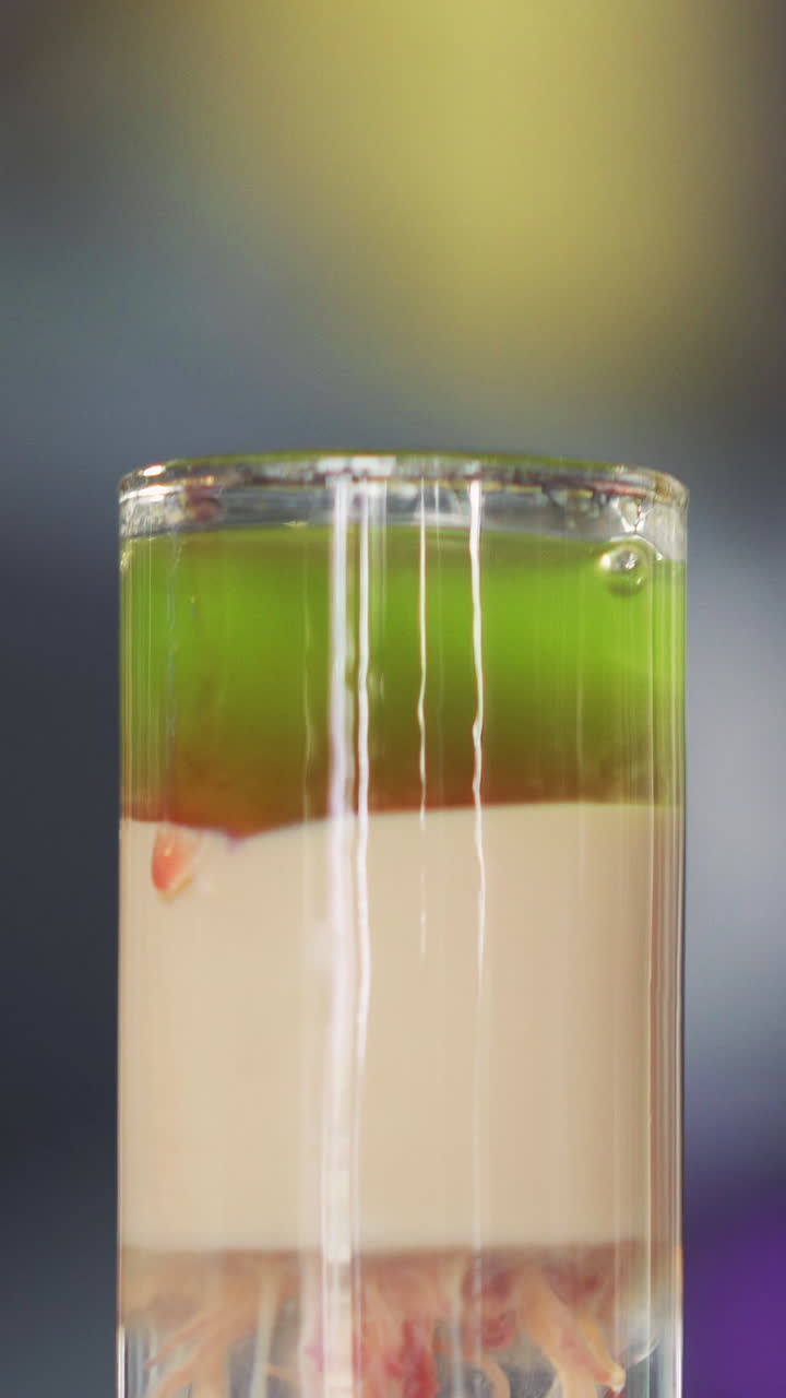 Barman adds red liquid into green and white cocktail into a glass on table. Red drops of alcohol falling in the cocktail. Vertical video.