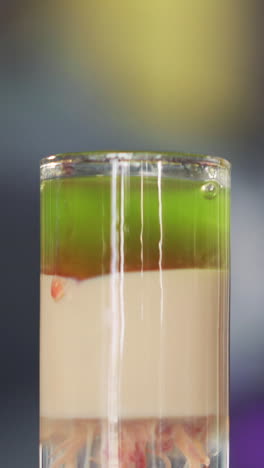 barman adds red liquid into green and white cocktail into a glass on table. red drops of alcohol falling in the cocktail. vertical video.