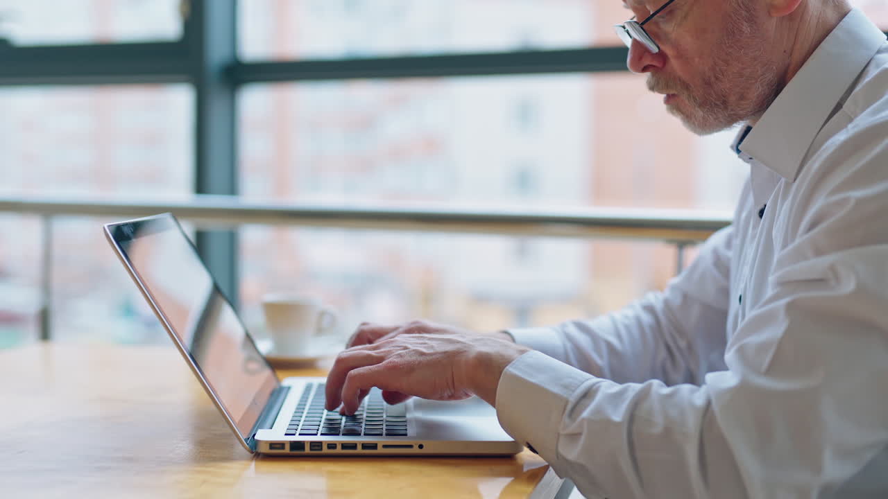 Senior businessman with laptop in office. Side view of a mature man finishing his work on a computer and taking a cup of coffee. Business concept. Close-up.
