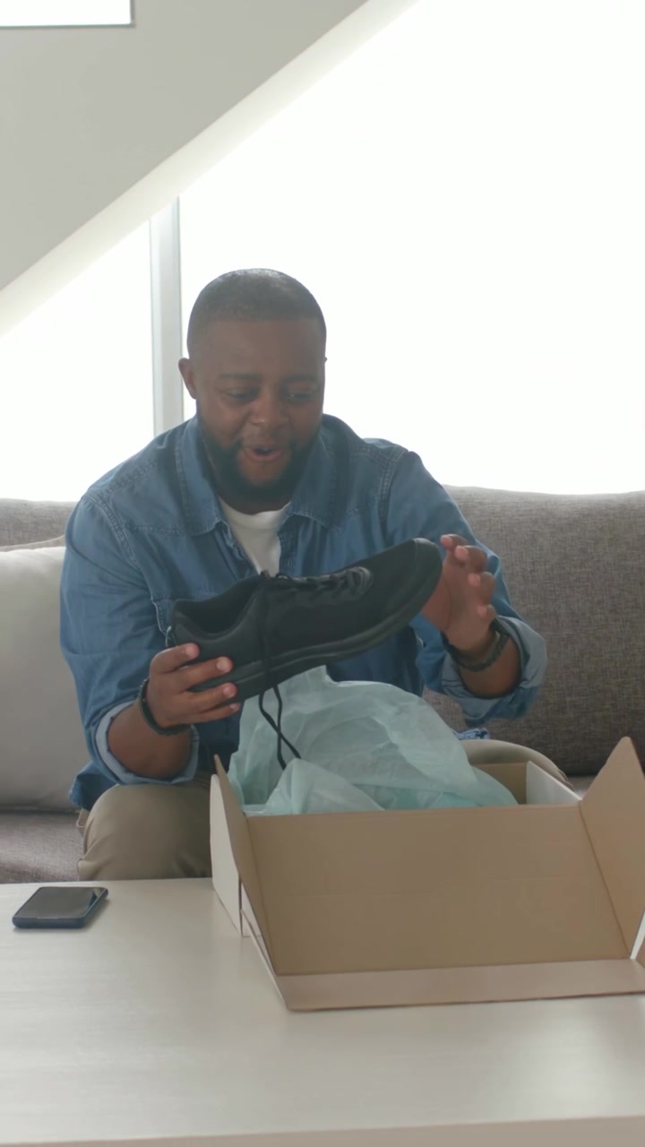 Vertical video: Opening box, man in denim checking black shoes on sofa, light-blue tissue showing