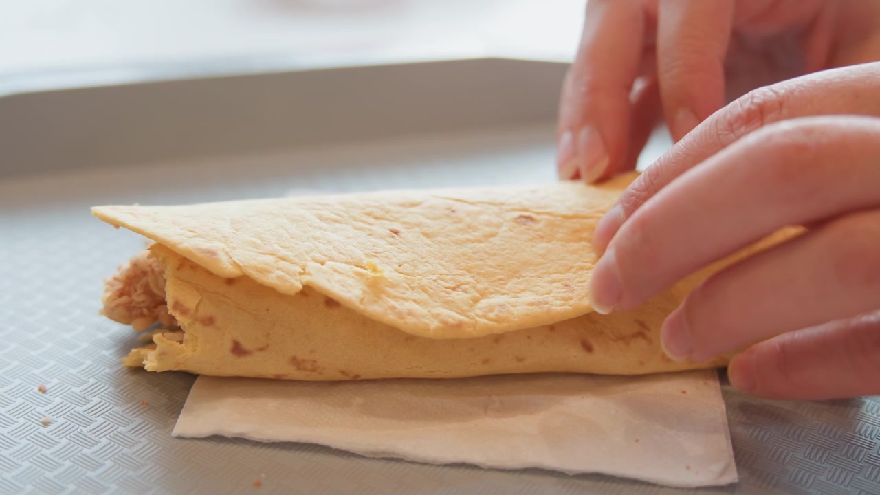 Vista cenital cercana muestra manos presionando pan plano delgado alrededor de tiras de pollo empanizadas crujientes, rodajas de pepinillo y salsa, formando un wrap para llevar durante la preparación casual en la cocina bajo una luz natural suave